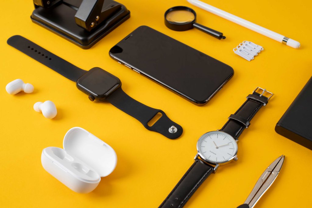 Flat lay of modern gadgets and accessories on a yellow background, including a smartphone, smartwatch, wristwatch, wireless earbuds, stylus pen, magnifying glass, and electronic tools.