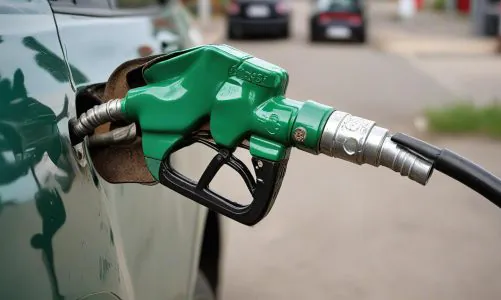 A green gas pump nozzle inserted into the fuel tank of a car, with other vehicles visible in the background on a street.