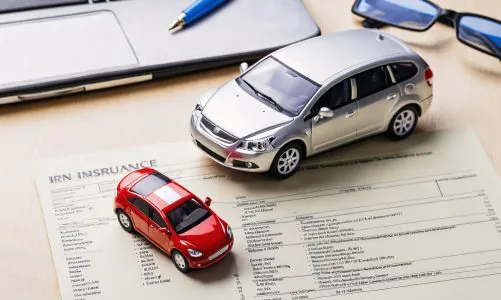Car insurance form with toy cars, pen, and laptop on a desk.