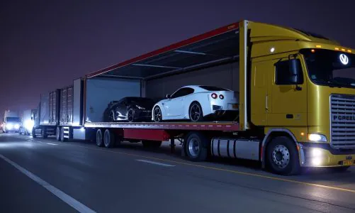 Luxury sports cars, including a white and a black model, being transported inside a yellow enclosed car carrier truck at night.