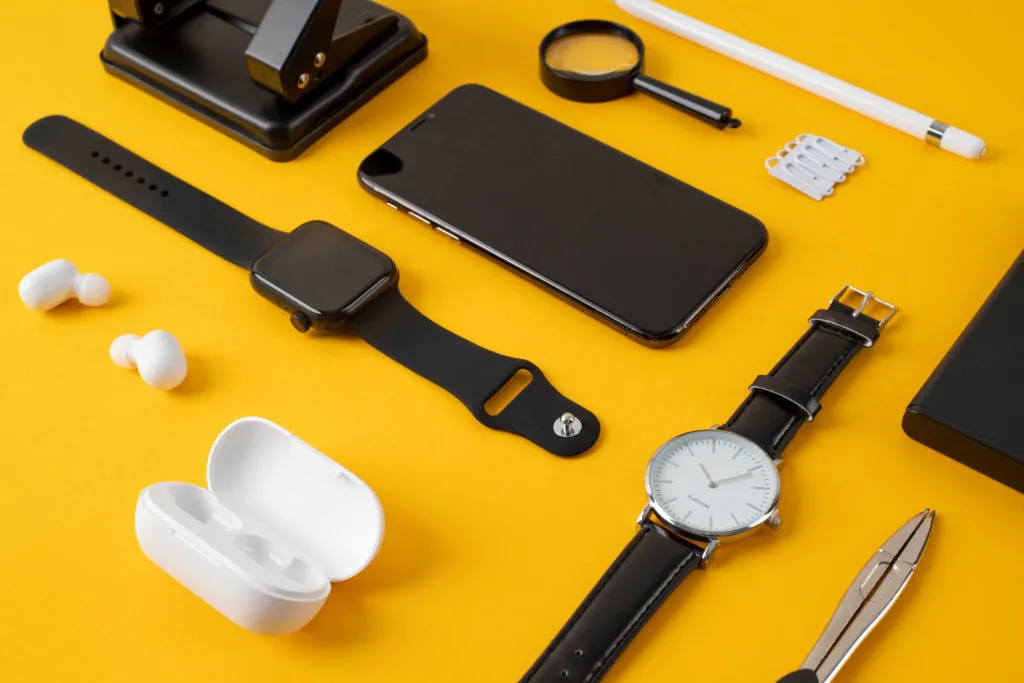 Flat lay of modern gadgets and accessories on a yellow background, including a smartphone, smartwatch, wristwatch, wireless earbuds, stylus pen, magnifying glass, and electronic tools.