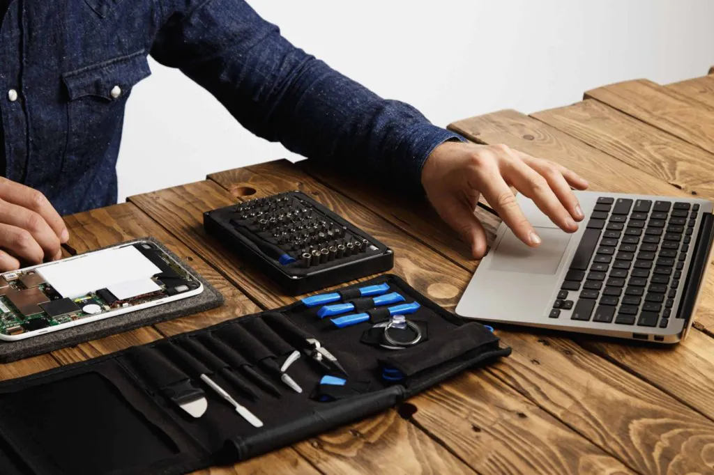 Technician working on electronic device repair at a wooden desk, with disassembled tablet parts, precision tools, and a laptop for diagnostics.