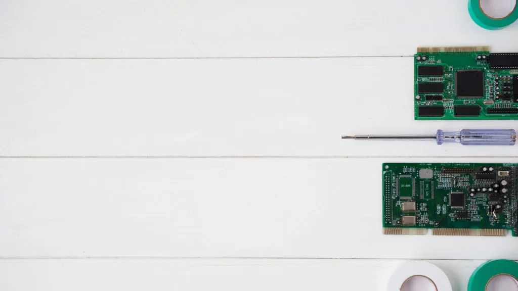 Top view of two green circuit boards and a screwdriver arranged neatly on a white wooden surface, with rolls of tape visible in the corners.
